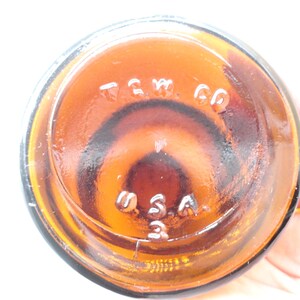 May include: The circular base of an amber-colored glass bottle featuring embossed lettering that reads T.G.W. CO and U.S.A. 3. The glass is translucent and shows a dark brown hue with a smooth, reflective surface texture throughout the bottom.