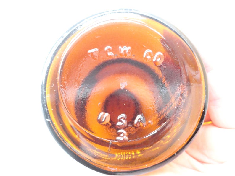 May include: The circular base of an amber-colored glass bottle featuring embossed lettering that reads T.G.W. CO and U.S.A. 3. The glass is translucent and shows a dark brown hue with a smooth, reflective surface texture throughout the bottom.