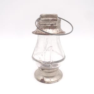 Pu&ograve; includere: Una piccola lanterna di vetro trasparente con parte superiore e base in metallo. La lanterna ha un manico e una forma decorativa. Le parti metalliche mostrano segni di et&agrave; e usura, con un aspetto arrugginito. La lanterna &egrave; progettata per contenere una piccola fonte di luce.