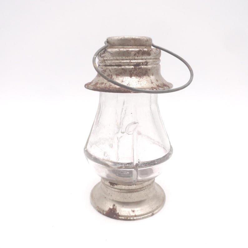 Pu&ograve; includere: Una piccola lanterna di vetro trasparente con parte superiore e base in metallo. La lanterna ha un manico e una forma decorativa. Le parti metalliche mostrano segni di et&agrave; e usura, con un aspetto arrugginito. La lanterna &egrave; progettata per contenere una piccola fonte di luce.