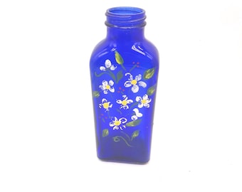Peut inclure: Une bouteille en verre bleu cobalt avec un goulot étroit et une base carrée. L'avant de la bouteille est peint à la main avec un motif floral composé de pétales blancs, de centres jaunes, de feuilles vertes et de petits points rouges.