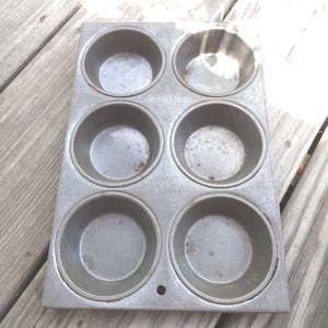 May include: A vintage metal muffin tin with six individual round molds. The tin is gray and shows signs of wear and use.