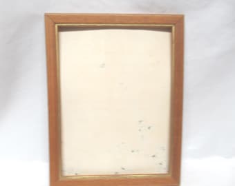 May include: A rectangular wooden picture frame with a light brown finish. The frame has a thin gold-colored inner border. The interior of the frame is a light beige color, with some dark spots.