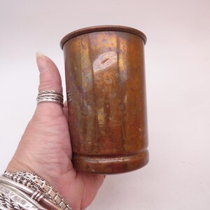 May include: A cylindrical copper cup with a weathered, oxidized patina featuring mottled brown and orange tones. The vessel has a smooth, straight-walled body with a slightly flared rim and a reinforced base, showing signs of age and use.