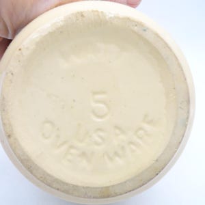 May include: Cream-colored ceramic ovenware with the number "5" and the text "U.S.A. OVEN WARE" embossed on the bottom. The piece has a rounded shape and a slightly textured surface.