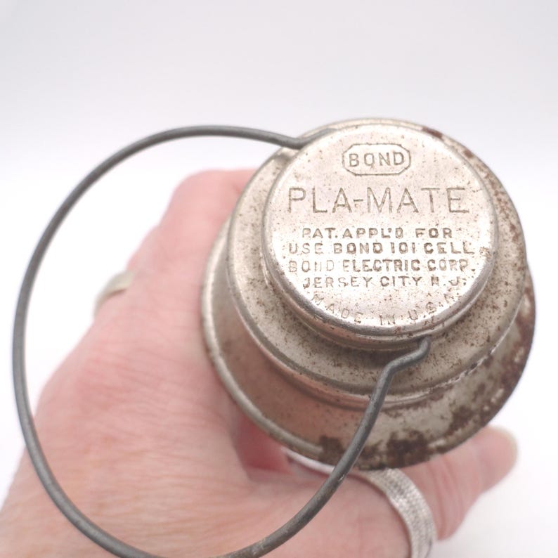 Pu&ograve; includere: Un contenitore in metallo vintage con la scritta "PLA-MATE" e "BOND" stampata sul coperchio. Il contenitore ha un manico in filo metallico ed &egrave; prodotto negli Stati Uniti. Il coperchio include anche il testo "PAT. APPLD FOR USE BOND 101 CELL BOND ELECTRIC CORP. JERSEY CITY N.J."
