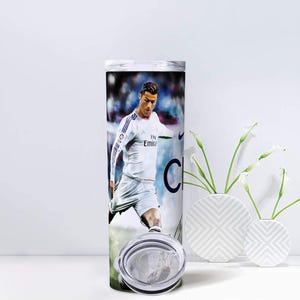 Puede incluir: Un vaso de acero inoxidable con una imagen a color de un futbolista con uniforme blanco. El dise&ntilde;o envuelve el cuerpo cil&iacute;ndrico del vaso, que incluye una tapa transparente. El fondo es una imagen borrosa de un campo de f&uacute;tbol.