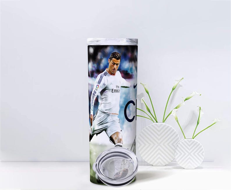 Puede incluir: Un vaso de acero inoxidable con una imagen a color de un futbolista con uniforme blanco. El dise&ntilde;o envuelve el cuerpo cil&iacute;ndrico del vaso, que incluye una tapa transparente. El fondo es una imagen borrosa de un campo de f&uacute;tbol.