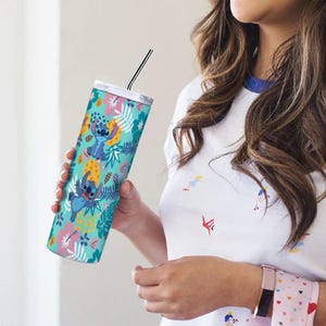 May include: A teal tumbler featuring the character Stitch from Lilo & Stitch, surrounded by a tropical floral pattern. The tumbler has a silver lid and a black and silver striped straw. The design includes blue, yellow, pink, and white elements.