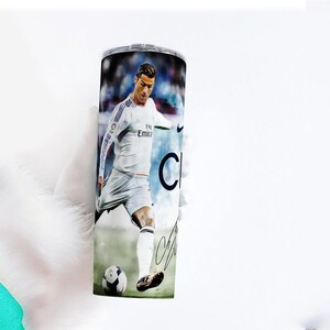 Puede incluir: Un vaso de acero inoxidable con una imagen a color de un futbolista con uniforme blanco pateando un bal&oacute;n de f&uacute;tbol. El vaso tiene una tapa plateada y una firma negra. El fondo es un estadio borroso.