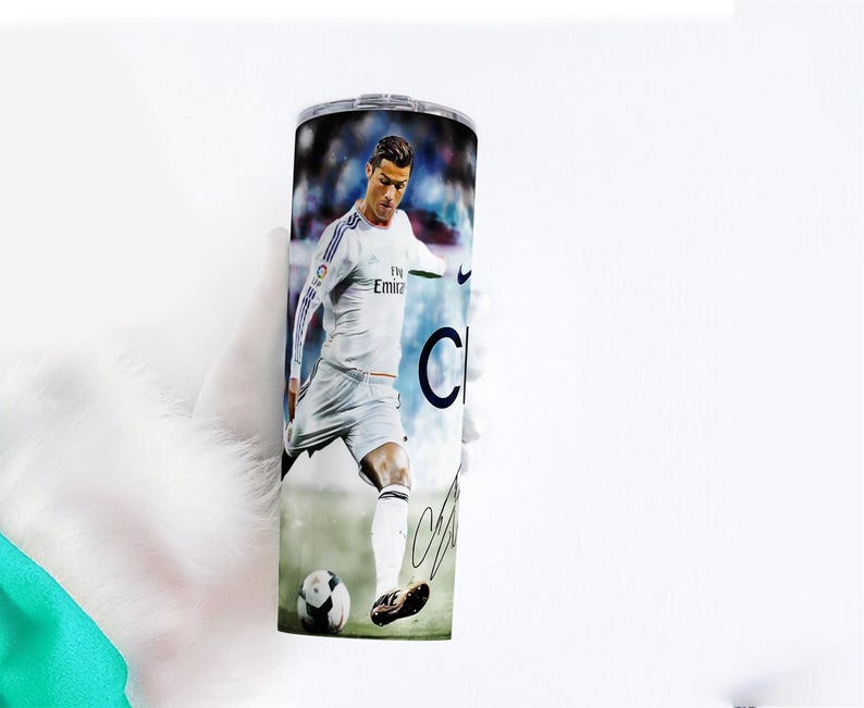 Puede incluir: Un vaso de acero inoxidable con una imagen a color de un futbolista con uniforme blanco pateando un bal&oacute;n de f&uacute;tbol. El vaso tiene una tapa plateada y una firma negra. El fondo es un estadio borroso.