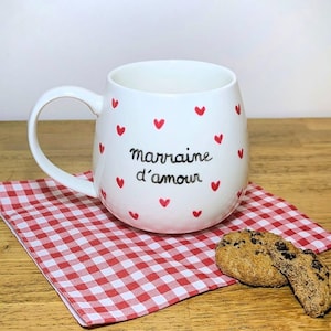 Peut inclure: Une tasse en céramique blanche de forme arrondie, ornée de petits cœurs rouges et du texte noir "marraine d'amour" imprimé sur le devant. La tasse possède une anse et est posée sur une serviette à carreaux rouges et blancs près d'un biscuit.