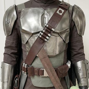 May include: A costume featuring a metallic silver chest plate, shoulder pauldrons, and gauntlets worn over a dark brown fabric suit. A brown leather bandolier with silver cartridges crosses the chest, paired with a matching utility belt.