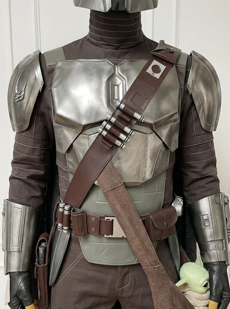 May include: A costume featuring a metallic silver chest plate, shoulder pauldrons, and gauntlets worn over a dark brown fabric suit. A brown leather bandolier with silver cartridges crosses the chest, paired with a matching utility belt.