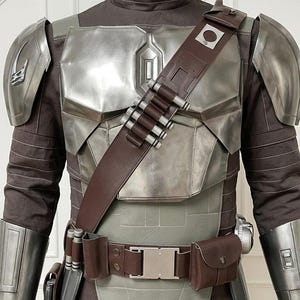 May include: A costume set featuring a dark brown long-sleeved shirt with a high collar, metallic silver chest and shoulder armor, and a brown leather bandolier with silver cylindrical cartridges. A matching brown utility belt includes pouches.