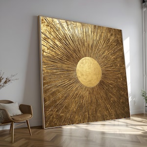 Peut inclure: Grande œuvre d'art carrée dorée avec un motif de soleil. L'œuvre présente une surface texturée avec un élément circulaire central et des lignes rayonnantes. Encadrée dans un cadre fin et clair, elle est appuyée contre un mur blanc.