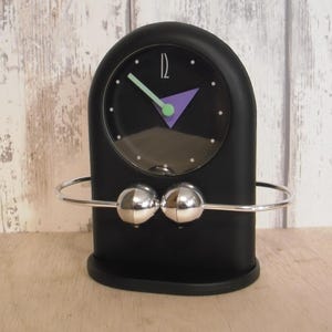 Puede incluir: Un reloj de escritorio negro con una esfera redonda, con un triángulo morado y manecillas verdes. El reloj tiene una barra de metal plateado con dos esferas plateadas. El reloj está sobre una base negra.
