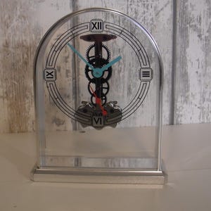 Puede incluir: Un reloj transparente en forma de arco con base y marco plateados. La esfera presenta números romanos y un mecanismo de engranajes. Las manecillas son de color azul claro. Es un objeto decorativo.