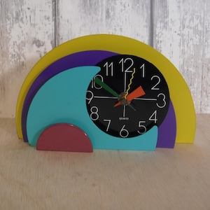 Puede incluir: Un reloj de cuarzo de estilo retro colorido con esfera negra y números blancos. El reloj presenta un diseño geométrico amarillo, morado, turquesa y rojo. Las manecillas son verdes, naranjas y amarillas. El reloj mide unos 18 cm de ancho.