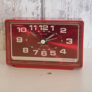 May include: A red rectangular alarm clock with white numerals and hands. The clock face has the text "RHYTHM" at the bottom. The clock is set against a light-colored background.