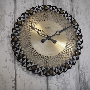 May include: A round, vintage wall clock with a gold-toned face and black geometric detailing. The clock features black hands and a decorative black outer ring with triangular cutouts. The clock is mounted on a weathered, light-colored wooden wall.