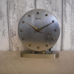 May include: A vintage silver-toned desk clock with a round face and gold-colored hands and hour markers. The clock has the name "Roma" printed on the face and sits on a rectangular base. The clock is a classic piece of decor.