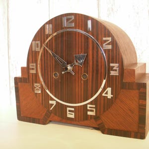 May include: A vintage wooden clock with a dark brown, striped face and a silver-coloured circular frame. The clock has silver numerals and hands. The clock's body is a dark brown wood with a geometric design.