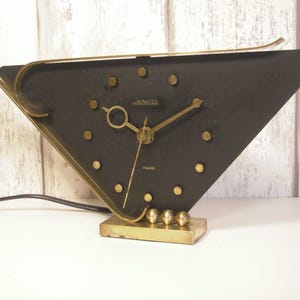 May include: A vintage black and gold-toned clock with a triangular face. The clock features gold-colored hands, hour markers, and a decorative frame. The base is rectangular with three gold spheres. The clock has a power cord.