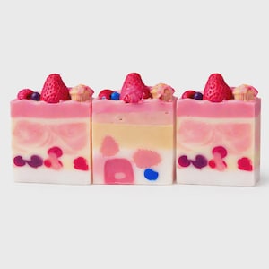 May include: Three decorative soap bars, designed to look like cake slices, are displayed. Each bar has layers of pink, white, and yellow, and is topped with a strawberry, berries, and miniature cupcake decorations. The soap has a dessert theme.