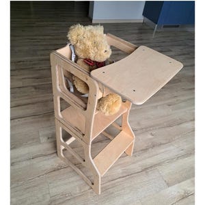 Peut inclure: Une tour d'apprentissage en bois clair avec une petite table et des sangles de sécurité. Un ours en peluche avec un nœud papillon rouge est assis à l'intérieur. La tour est conçue pour les enfants.
