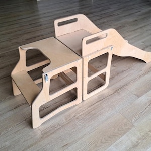 Op de afbeelding: Een houten speelset met een glijbaan en twee opstapblokken. De set is gemaakt van lichtgekleurd hout en is ontworpen voor kinderen. De glijbaan is bevestigd aan een van de blokken en het andere blok heeft een uitsparing om op te klimmen.