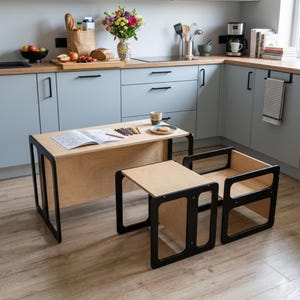 Puede incluir: Un juego de mesa y silla para niños de madera clara con marcos negros. La mesa tiene un libro para colorear y lápices de colores. La silla es un diseño de caja pequeña y abierta. El conjunto está en una cocina.