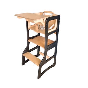 May include: A wooden learning tower with a black frame and natural wood shelves. The tower has a small table, a safety harness, and a handle. It is designed to help children reach countertops and participate in activities.
