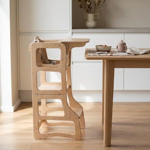 Puede incluir: Una torre de aprendizaje de color madera natural con una barra de seguridad y reposapiés. La torre tiene un diseño rectangular con aberturas recortadas y está colocada junto a una mesa de madera. El entorno de la cocina incluye un cuenco, una taza y un jarrón.
