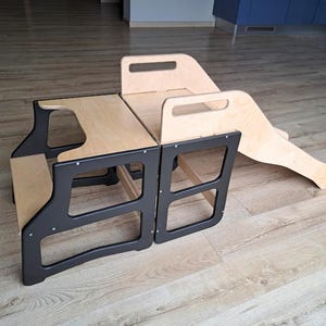 Könnte beinhalten: Ein hölzernes Kleinkind-Trittschemel- und Rutschbahn-Set. Das Set besteht aus zwei schwarz gerahmten Hockern mit Oberflächen aus Naturholz und einer passenden Rutsche. Die Hocker haben ausgeschnittene Griffe und sind für einfache Bewegung und Nutzung konzipiert.