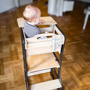 May include: A wooden high chair with a black frame. The chair has a tray, a seat, and a footrest. A baby wearing a striped top is seated in the chair. The chair has a safety harness.