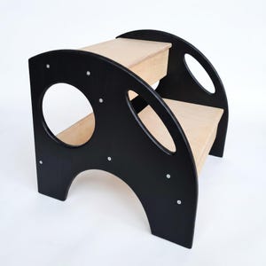May include: A two-step wooden step stool with a black frame and natural wood steps. The frame has circular and oval cutouts. The step stool is designed for children and has a sturdy construction.