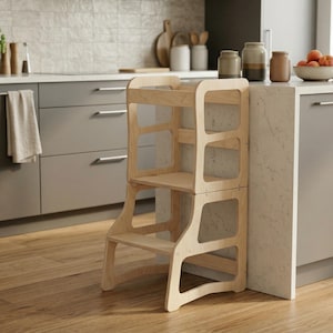 Puede incluir: Un taburete de cocina de madera clara con un diseño de dos escalones. El taburete tiene un marco rectangular con bordes redondeados y secciones recortadas. Está diseñado para ayudar a los niños a alcanzar encimeras y otras superficies elevadas.