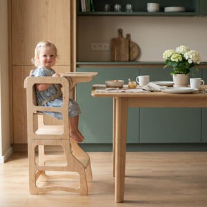 Könnte beinhalten: Ein Lernturm aus hellem Holz mit integriertem Tablett und Sicherheitsgurt. Die Struktur zeichnet sich durch ein hohes, offenes Rahmendesign mit einer Plattform zum Stehen oder Sitzen aus, gefertigt aus glattem, natürlichem Holz.