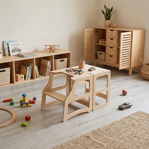 May include: A light-coloured wooden learning tower with a step stool design, featuring a convertible structure with metal hinges. The tower is positioned in a room with a wooden storage cabinet, a low bookshelf, and various scattered children's toys.