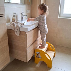 Peut inclure: Un marchepied en bois jaune à deux marches, conçu pour aider un enfant à atteindre un lavabo de salle de bain. Le tabouret a des motifs découpés sur les côtés et est placé devant un évier blanc. Un enfant se tient sur le tabouret, tendant la main vers l'eau courante.