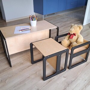 May include: A child's furniture set featuring a light wood table and two smaller tables with black frames. A teddy bear with a red bow sits on one of the smaller tables. Colored pencils and paper are on the table.