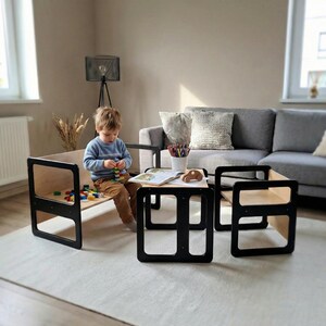 May include: A child's wooden table and chair set with a black frame and natural wood surfaces. The set includes a small table and two chairs, with a child sitting on one of the chairs. The child is playing with colorful blocks.