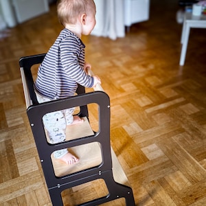May include: A black and natural wood toddler step stool with a child wearing a striped shirt and patterned pants. The step stool has a rectangular base and three openings. The child is standing on the top step, holding onto the side rails.
