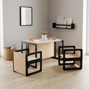 Puede incluir: Un moderno juego de mesa y sillas para niños. La mesa y las sillas tienen un acabado de madera natural con detalles en negro. El conjunto incluye una mesa rectangular, dos sillas cuadradas y una estantería de pared negra. La mesa mide unos 76 cm de largo.