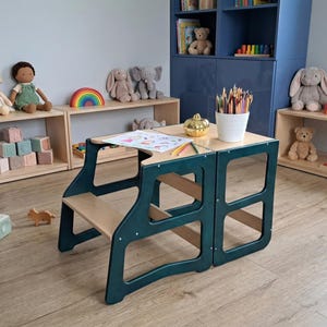 Puede incluir: Una torre de aprendizaje de madera clara y verde oscuro que se convierte en mesa y silla. La estructura presenta una superficie plana con un dibujo en papel de figuras coloridas, un vaso blanco lleno de lápices de colores y una calabaza dorada.