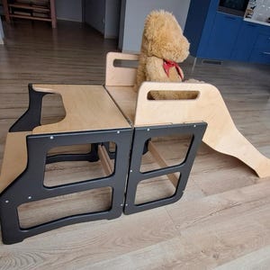 May include: A wooden children's play set with a slide and steps. The set is made of light-coloured wood with black accents. A stuffed teddy bear with a red bow sits on the play set.