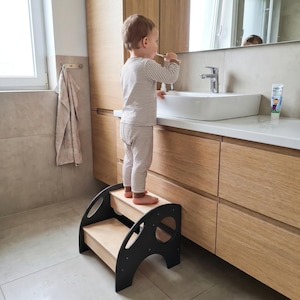 May include: A two-step wooden stool featuring a light wood finish on the steps and dark grey side panels with circular cutouts. The stool is designed with a sturdy frame and provides elevation for reaching the bathroom sink.