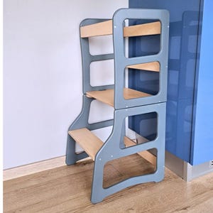 May include: A grey wooden learning tower with natural wood shelves and steps. The tower has a sturdy frame and is designed to help children reach countertops and other elevated surfaces. The tower is placed against a white wall and a blue cabinet.
