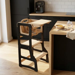 May include: A black and natural wood learning tower with a safety harness and a small table. The tower has two steps and a platform for a child to stand on. The tower is designed to help children reach countertops and other elevated surfaces.